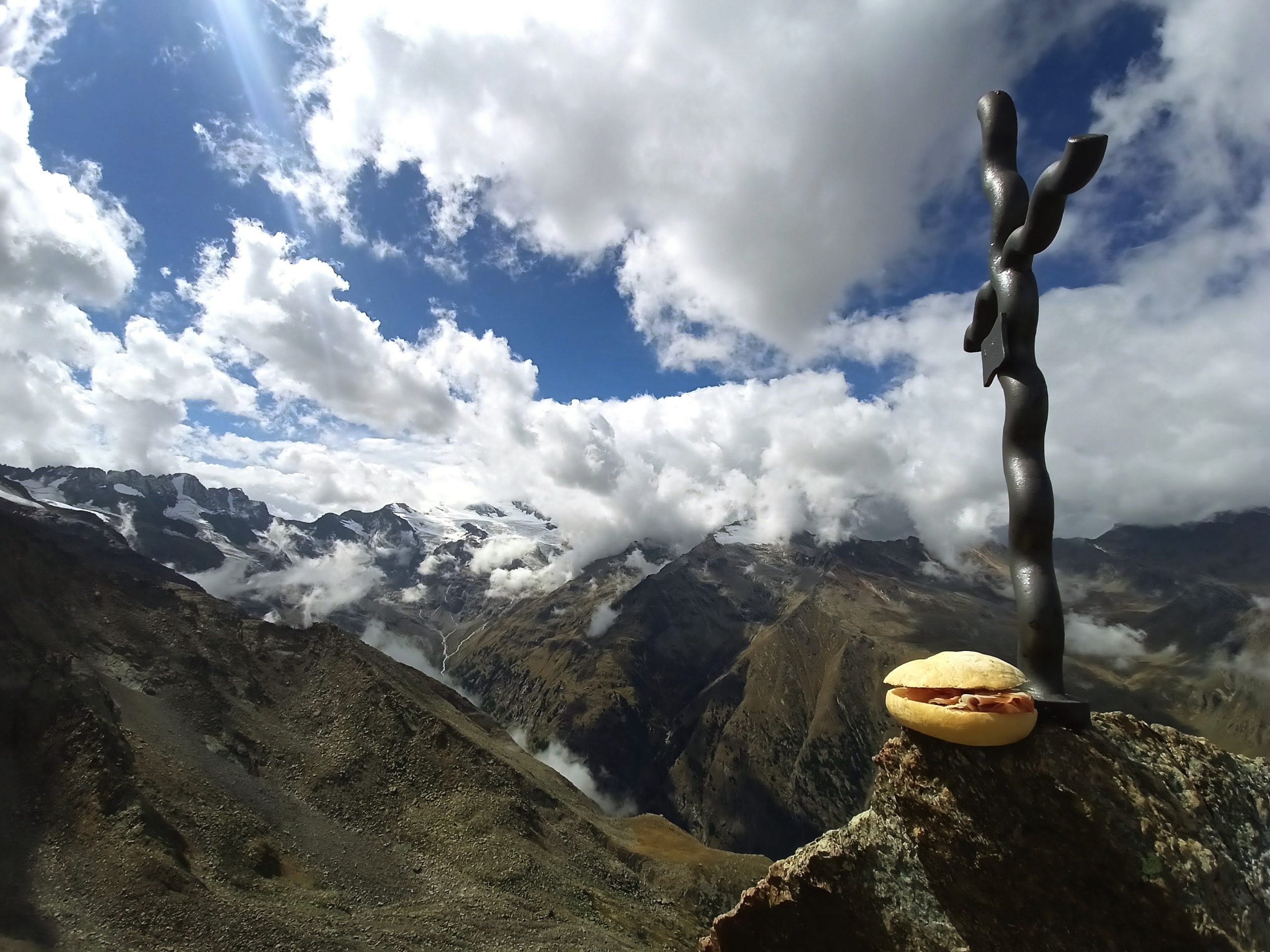 Un panino e una croce sulla vetta di una cima. Sullo sfondo imponenti nuvole bianche si stagliano nel cielo blu