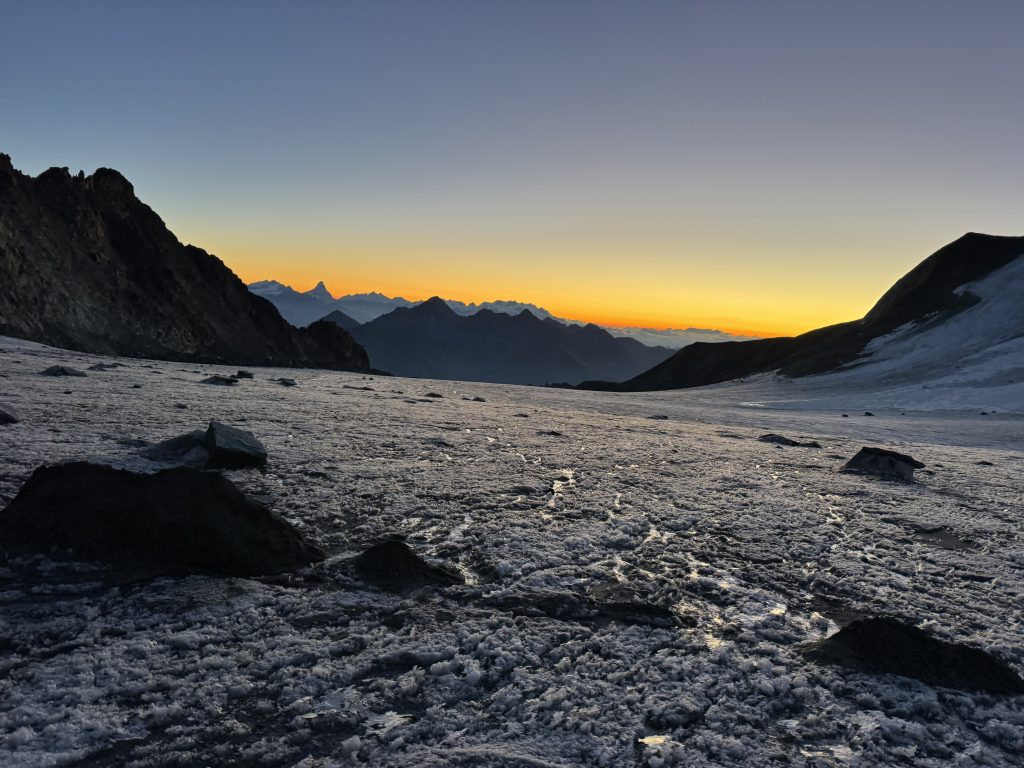 Un ghiacciaio cosparso di sassi in primo piano, sullo sfondo un'alba e delle alte montagne fanno capolino