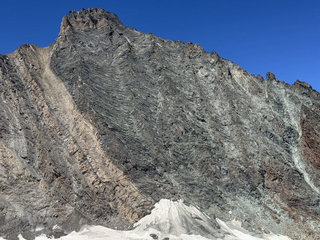 La parete rocciosa su cui si trova la via normale della Grivola è solcata da diversi speroni. Un cono di neve si trova alla sua base. Il cielo è blu.