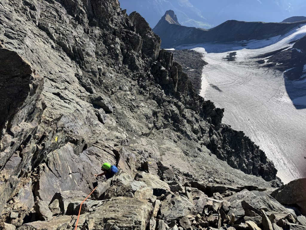 Sulla parete rocciosa della via normale della Grivola si vede un arrampicatore. Lo scalatore ha un casco verde e una maglia blu, la corda a cui è legato è arancione. Sullo sfondo si vede la larga distesa nevosa di un ghiacciaio