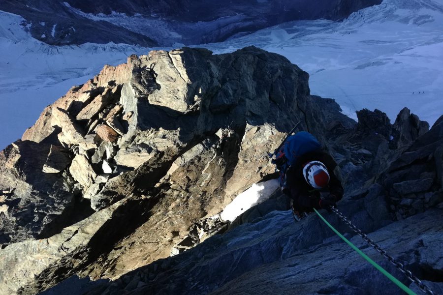 Una persona si arrampica su una ombrosa parete rocciosa del Polluce. La persona è assicurata da una corda verde. Sulla parte sinistra si vedono i primi raggi di sole sulla parete.