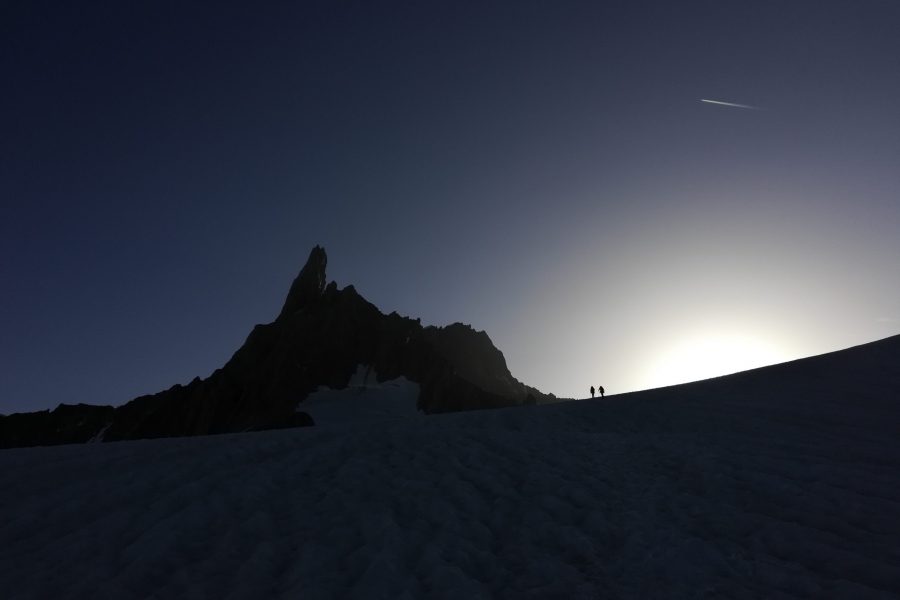 Le prime luci del giorno illuminano il cielo. In centro si erge ripido un pilastro di roccia denominato Dente del Gigante. In primo piano c'è un pendio di neve su cui, a fil di cielo, si vedono due persone che camminano.
