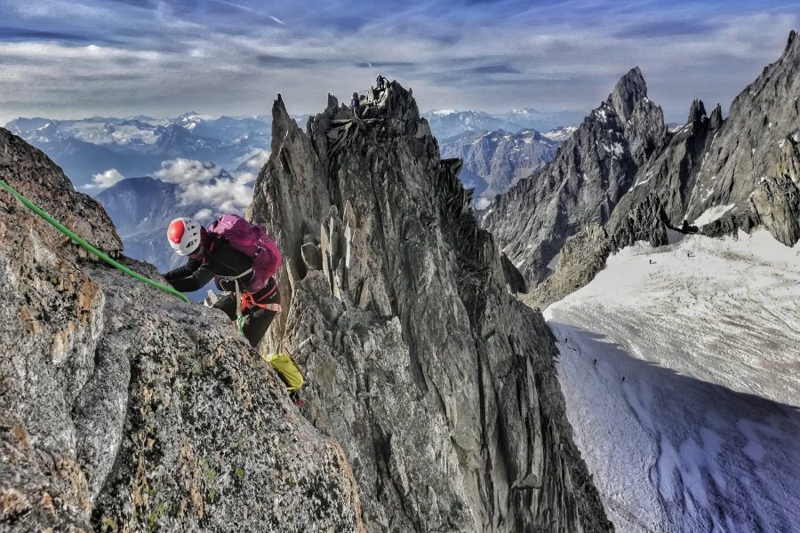 Una persona percorre un'affilata cresta rocciosa a fil di cielo. La persona è assicurata da una corda verde, ha un casco bianco e arancione e uno zaino viola. Sullo sfondo in basso a destra c'è un ghiacciaio. Il cielo ha qualche nuvola bianca.