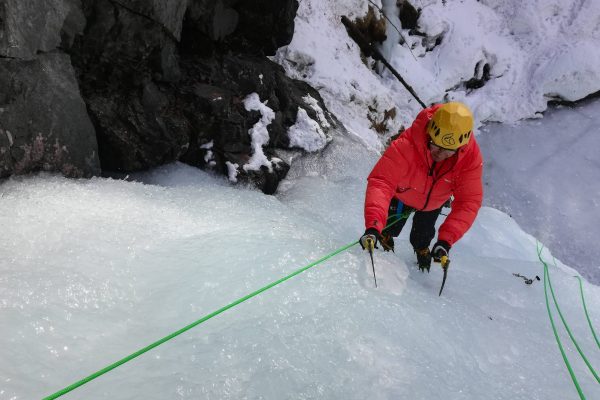 uno scalatore sale una cascata di ghiaccio assicurato da una corda verde. Egli ha una giacca rossa e un casco giallo. Sulla sinistra la cascata è circondata da una parete rocciosa