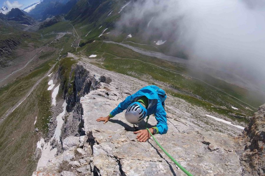 Una persona vestita di blu arrampica sulla cresta rocciosa delle Pyramides Calcaires. Sullo sfondo il fondo valle con dell'erba verde e una chiazza di neve. Una nuvola rende l'atmosfera molto suggestiva.